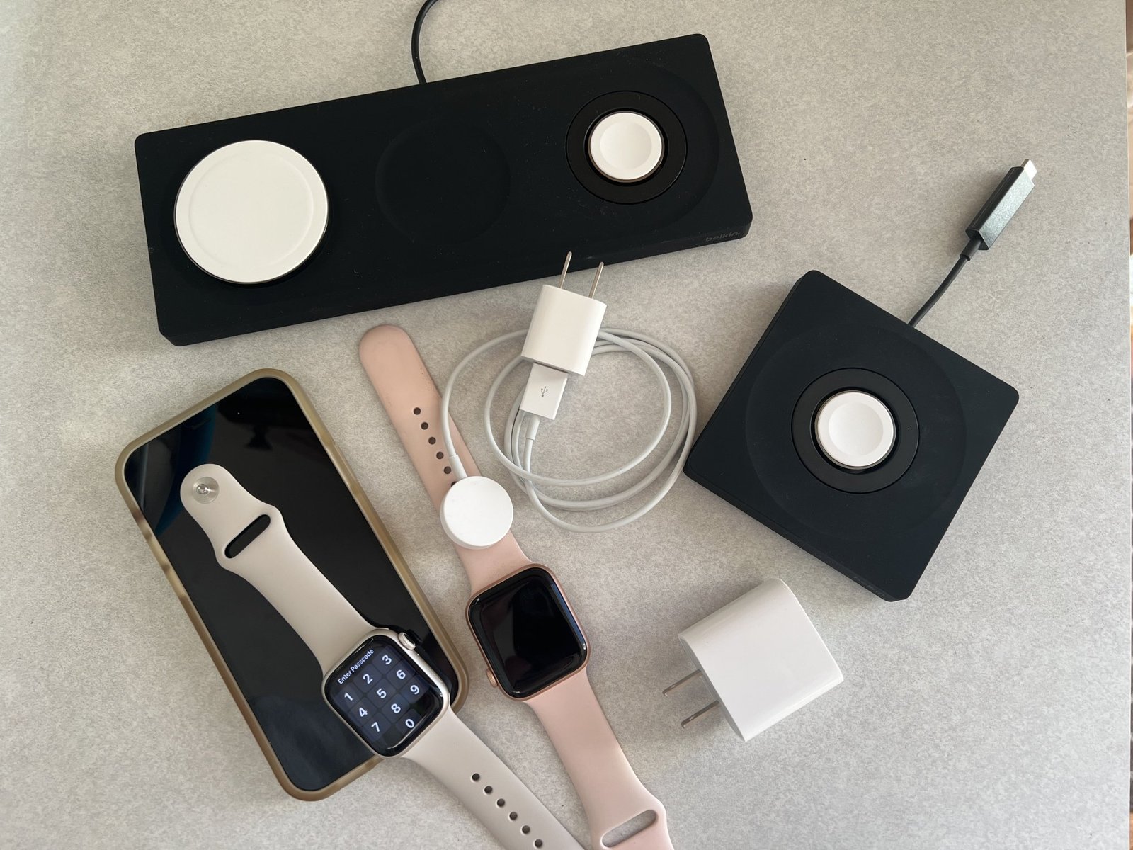 A collection of chargers, including the Belkin 3-in-1 pad, the Belkin Apple Watch Charger, the Apple Watch charger, a 20 watt USB-C adaptor, an iPhone 12, and two Apple Watches.