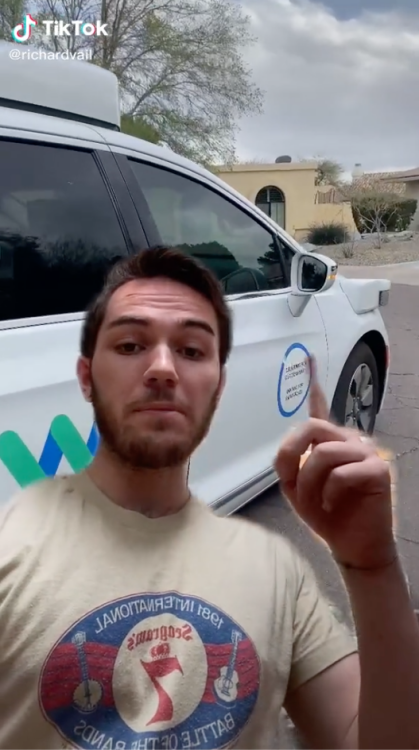 A man in a beige T-shirt standing in front of a white minivan. 